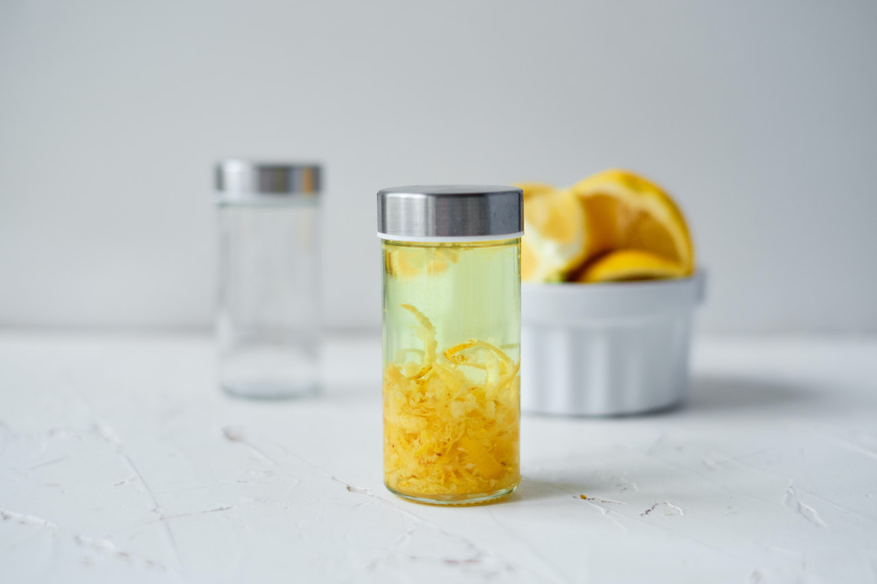 a glass jar filled with lemon peels and vodka, sealed. a bowl of lemon slices and a second empty jar in the background