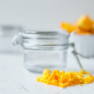 orange peels sitting on the counter in front of a small mason jar. Second jar and slices of oranges in a white bowl in the background