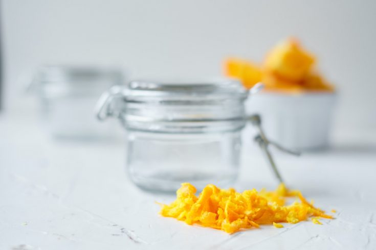 orange peels sitting on the counter in front of a small mason jar. Second jar and slices of oranges in a white bowl in the background