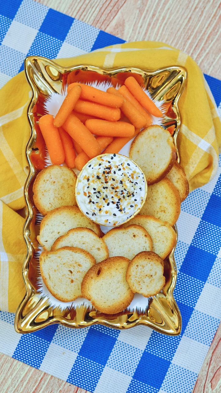 a bowl of everything but the bagel dip sitting on a gold platter surrounded by bagel chips and carrots