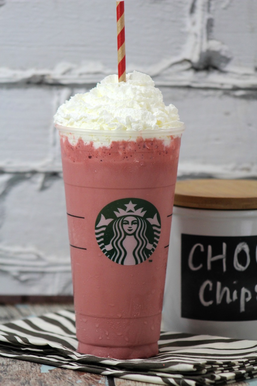 a clear tall glass with the Starbucks logo filled with pink Frappuccino topped with whipped cream with a red and white striped straw
