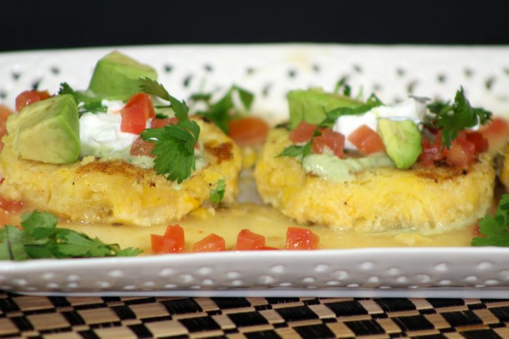 two tamale cakes sitting on a white plate topped with pico, sour cream, and avocado chunks