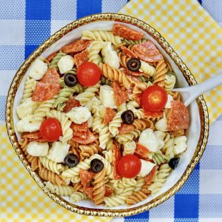 a gold rimmed large bowl filled with zesty Italian pasta salad. sitting on top of a yellow checkered and blue checkered towel and placemat