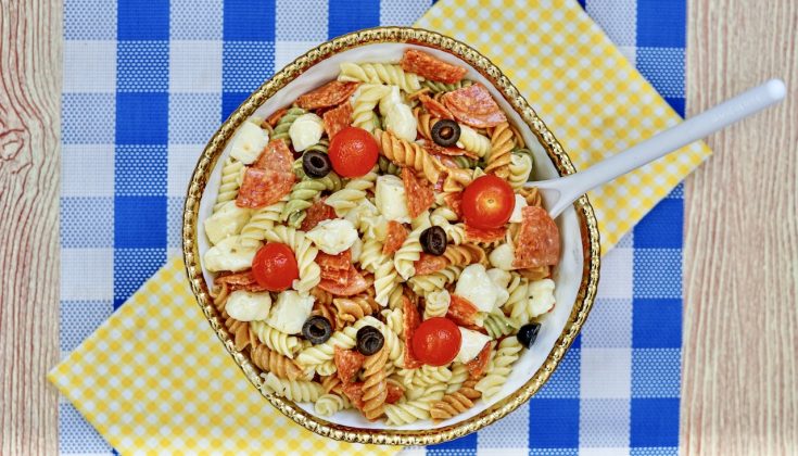 a gold rimmed large bowl filled with zesty Italian pasta salad. sitting on top of a yellow checkered and blue checkered towel and placemat