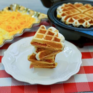 a stack of 4 stuffed waffles sitting on a round white plate on a red and white checkered placemat. Rectangular dish with scrambled eggs and shredded cheese to the left and an open waffle iron with another waffle to the right