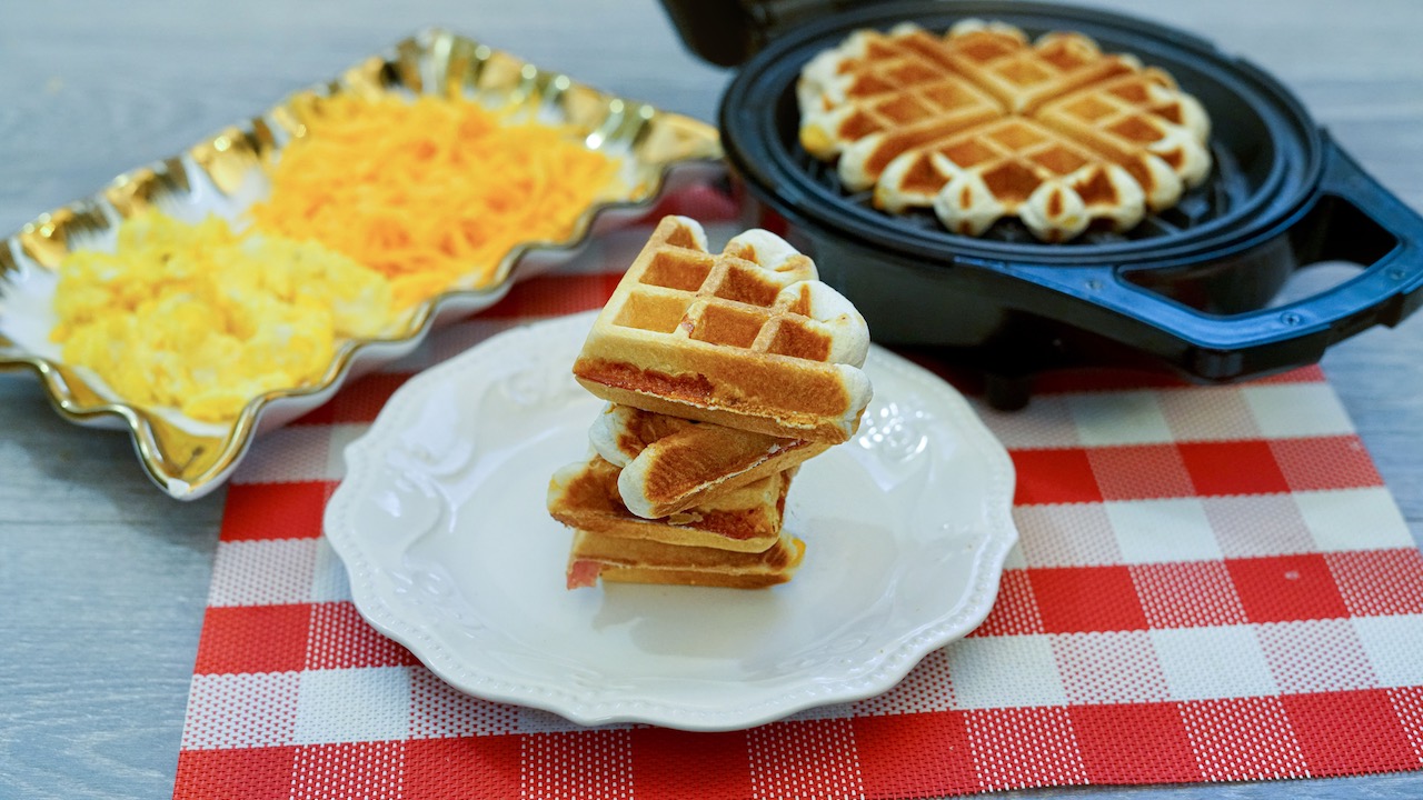 a stack of 4 stuffed waffles sitting on a round white plate on a red and white checkered placemat. Rectangular dish with scrambled eggs and shredded cheese to the left and an open waffle iron with another waffle to the right 
