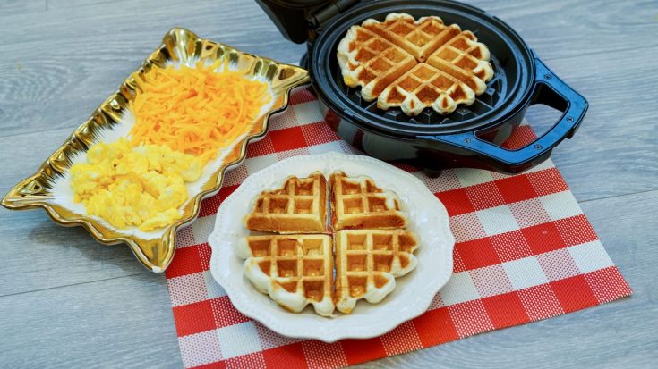 a white plate topped with a full stuffed waffle sitting on a red and white checkered place mat. A rectangular plate with shredded cheese and scrambled eggs to the left, a waffle iron with a second waffle to the right