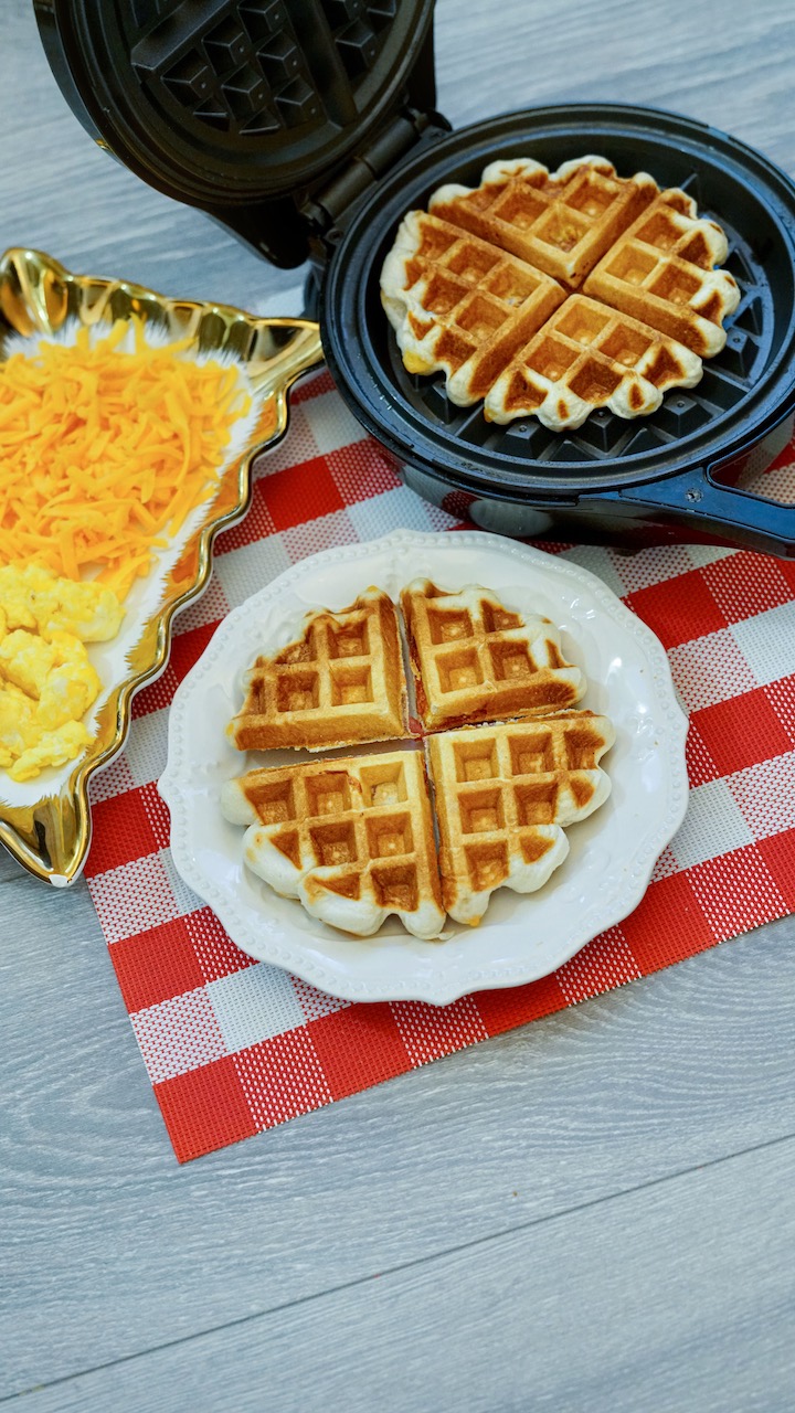 a white plate topped with a full stuffed waffle sitting on a red and white checkered place mat. A rectangular plate with shredded cheese and scrambled eggs to the left, a waffle iron with a second waffle to the right