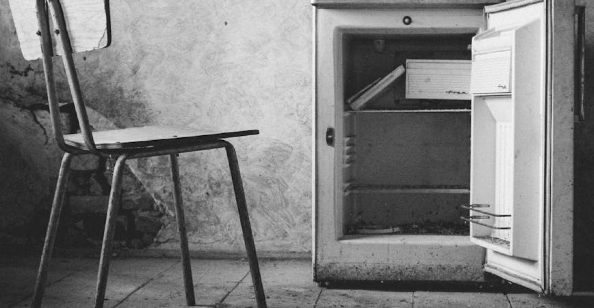 Black and white photo of an abandoned room with a chair and open mini fridge