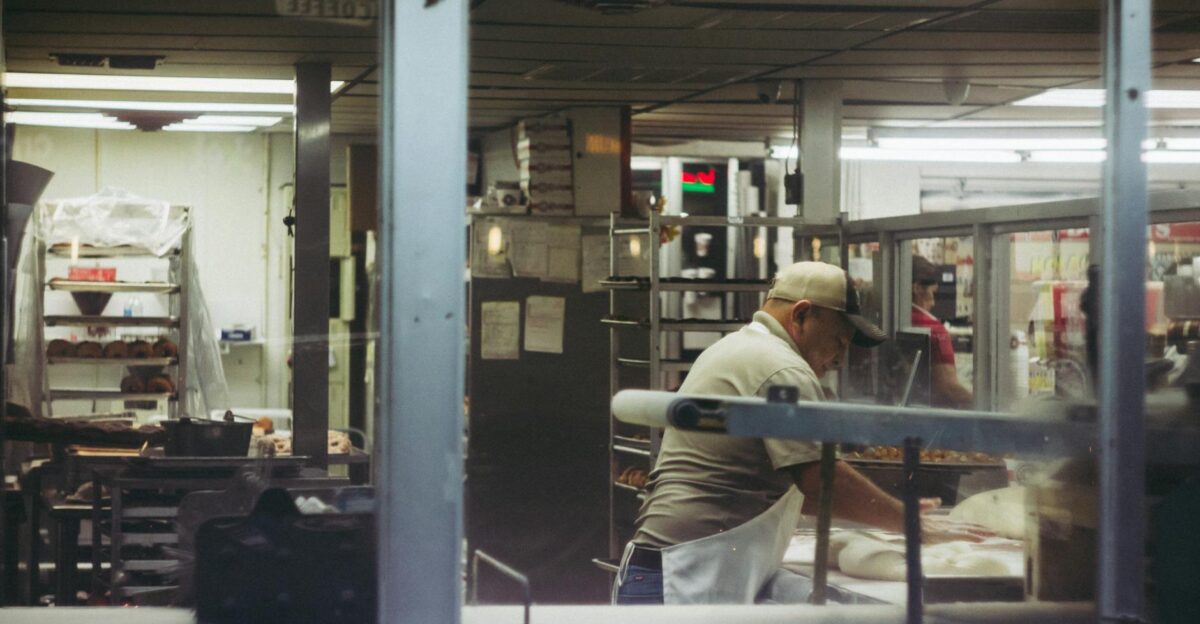 Baker working with dough in a commercial bakery kitchen viewed through a glass window