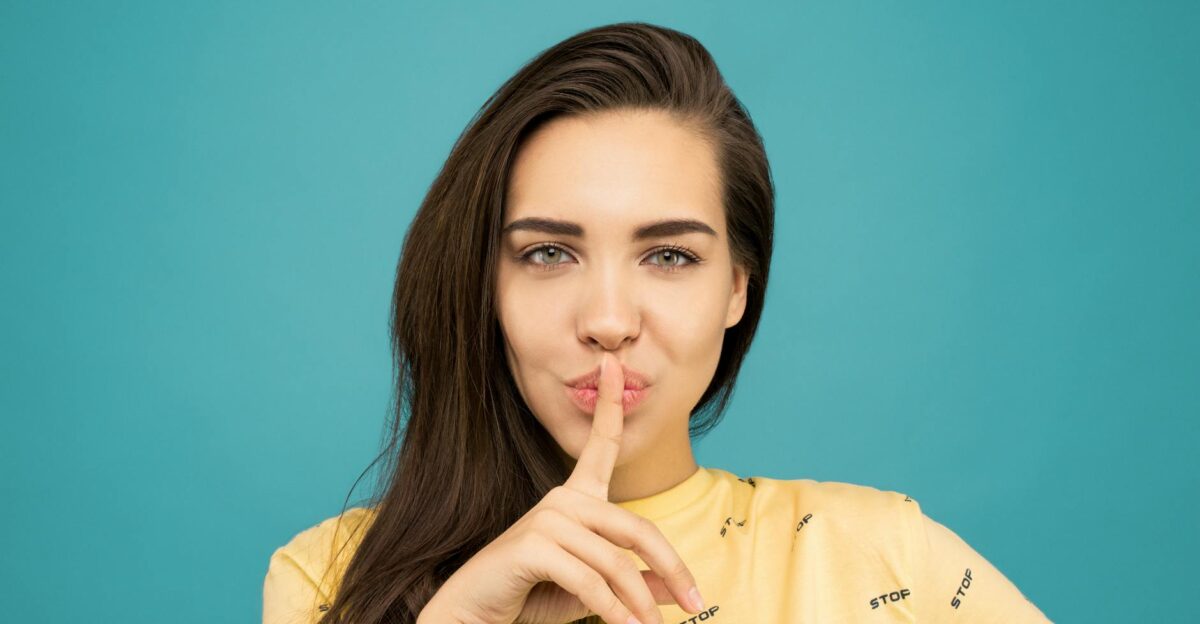 Portrait of a woman with brown hair posing with a hush gesture against a blue background