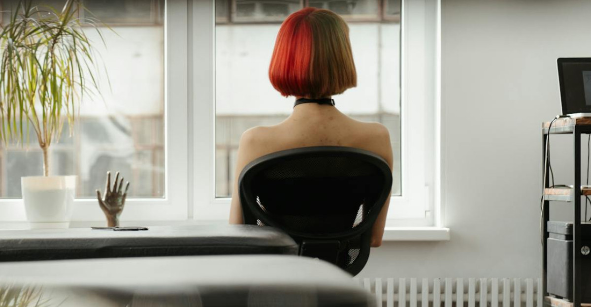 A woman with vivid red hair sits by the window in a modern sunlit tattoo studio