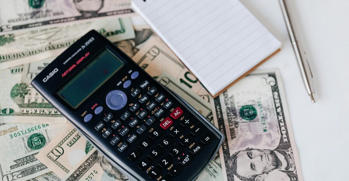 From above electronic calculator and notepad placed over United States dollar bills together with metallic pen for budget planning and calculation