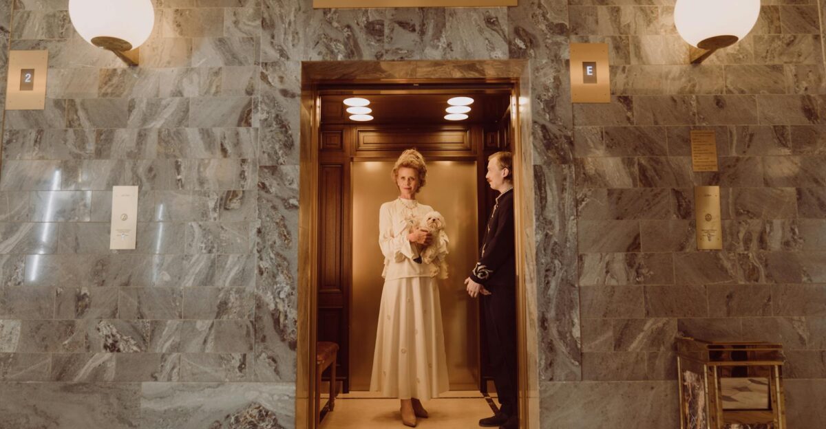 A refined lady with a small dog in a vintage hotel elevator accompanied by a doorman