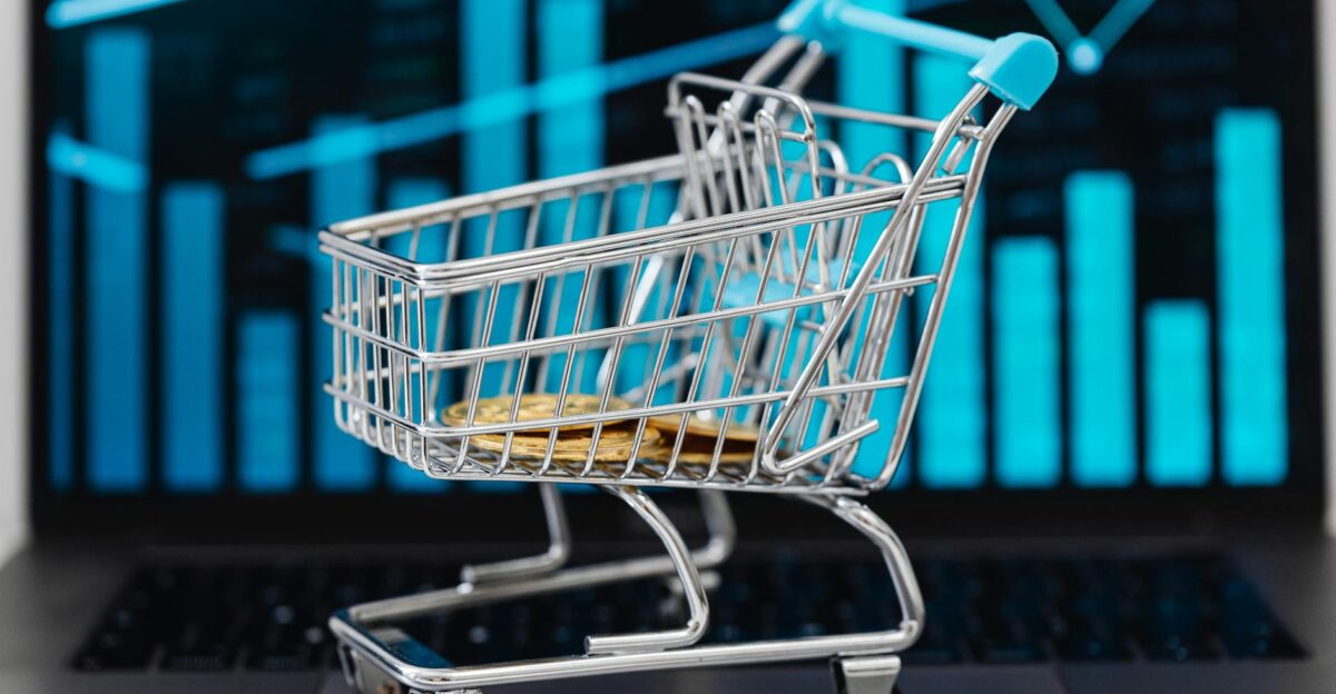 A miniature shopping cart with coins sits on a laptop displaying financial graphs symbolizing e-commerce and online shopping trends