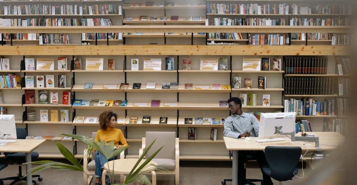 Two adults engaged in reading activities in a public library with abundant bookshelves