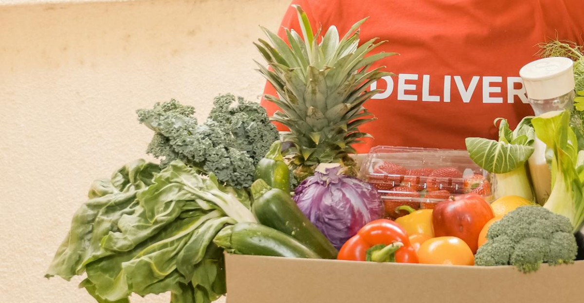 Deliveryman in red shirt holding a box of fresh vegetables smiling at the doorway