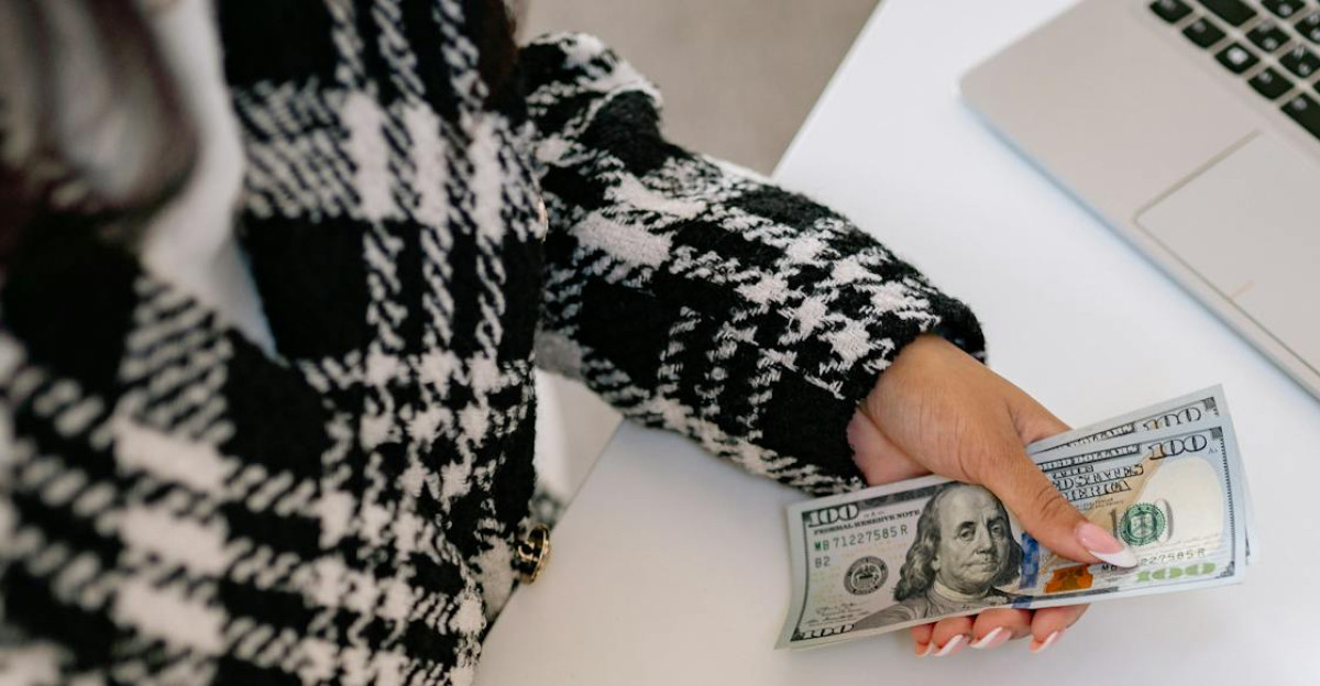 A woman in a plaid coat counts American dollars at a desk with a notebook and laptop