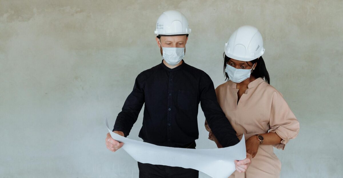 Two architects wearing face masks and hard hats review building plans indoors ensuring safety