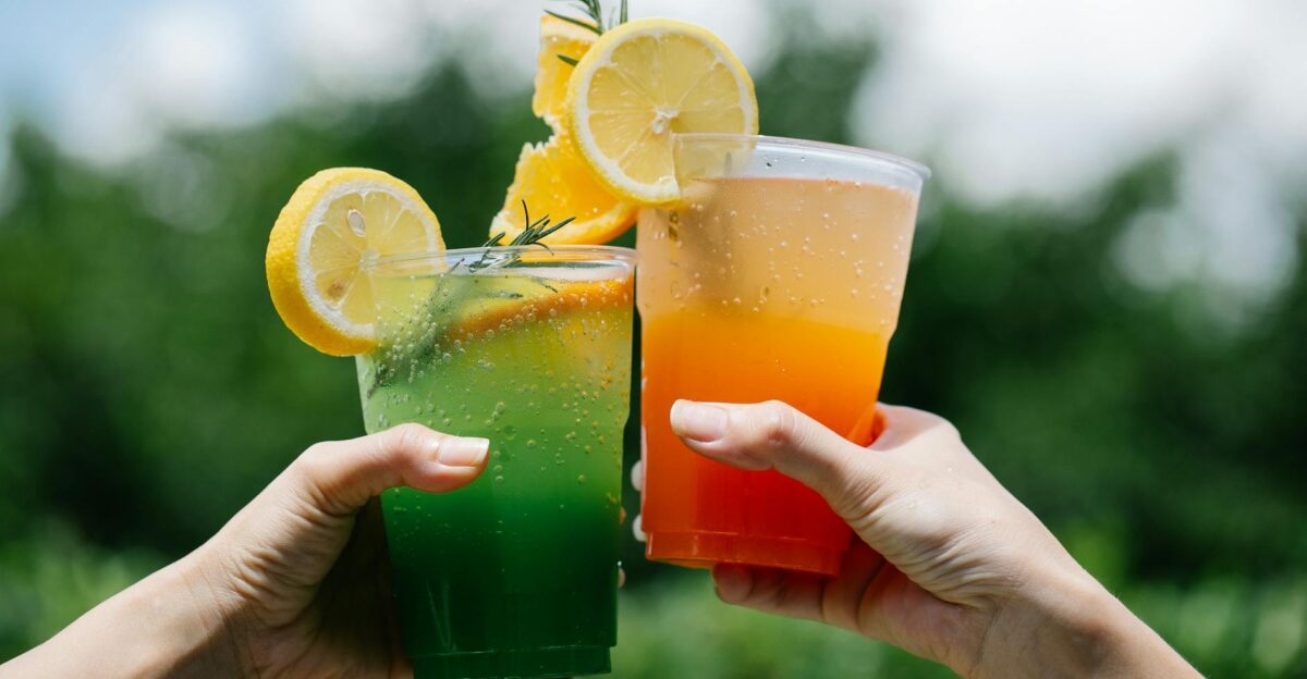 Two hands holding colorful citrus lemonades in a vibrant summer outdoor setting